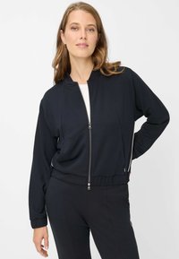 Schwarze Jacke mit Reißverschluss, geripptem Kragen und Bündchen, Seitenstreifen und leicht verkürztem Schnitt. Weicher Stoff mit glatter Textur.