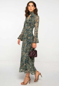 Mujer vistiendo un vestido largo con estampado y mangas transparentes, sosteniendo un pequeño bolso de mano púrpura oscuro, combinado con sandalias de tacón alto metálicas.