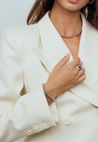 Kvinna i krämfärgad blazer som bär ett halsband med silverpärlor, matchande pärlarmband och en iögonfallande silverring, handen som försiktigt håller i slaget.
