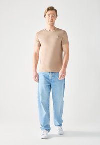 Camisa de manga corta beige con cuello redondo y pequeño emblema, combinada con unos vaqueros de mezclilla de corte holgado en azul claro y zapatillas blancas.