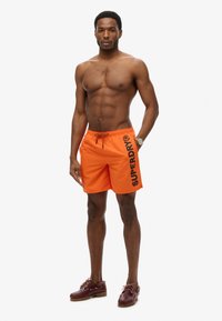Shorts de bain orange avec le logo "SUPERDRY" en noir. Tissu léger, taille élastique avec cordon de serrage. Portés avec des chaussures bateau marron.
