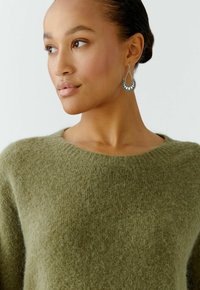 Olive green, soft wool sweater with a round neckline, paired with silver teardrop earrings featuring a sunburst design.