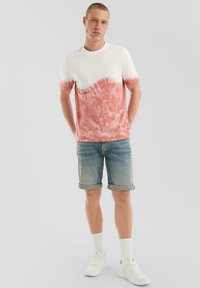 Witte en koraalkleurige tie-dye t-shirt met ronde hals, gecombineerd met opgerolde denim shorts. Model draagt witte sneakers en witte sokken.