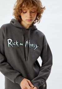 Harmaa huppari pehmeästä kankaasta, jossa on suuri, kontrastinen sininen ja musta "Rick and Morty" -grafiikka edessä.