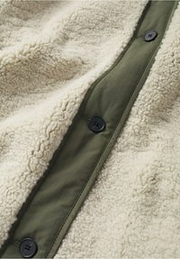 Gros plan d'une veste en tissu sherpa couleur crème avec une patte de boutonnage verte et de grands boutons noirs attachés.
