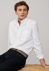 Camisa blanca de botones con un solo bolsillo en el pecho, combinada con jeans de mezclilla oscura y un cintur�ón trenzado beige, sentado sobre una superficie de madera.