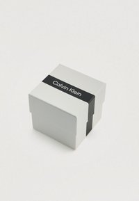 Boîte carrée blanche avec une bande noire portant l'inscription "calvin klein" au centre. Surface lisse, coins repliés, design minimaliste.