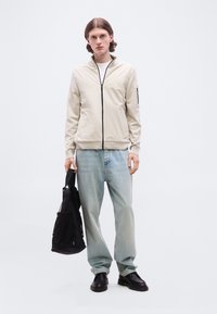 Νεαρός άνδρας με μεσαίου μήκους μαλλιά, φορώντας μπεζ ζακέτα με φερμουάρ, ανοιχτόχρωμο μπλε τζιν, μαύρα παπούτσια, κρατώντας μαύρη τσάντα στο αριστερό χέρι, στέκεται μπροστά σε ουδέτερο φόντο.