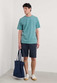 Turkos t-shirt med kort ärm, marinblå shorts, vita sneakers med bruna sulor, håller en marinblå tygpåse, står på ett ljust golv.