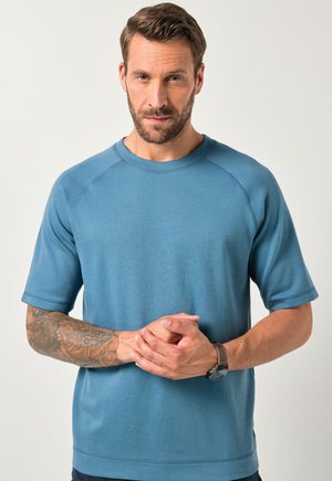 Blauwe T-shirt met korte mouwen, gemaakt van zachte stof. Heeft een ronde hals en raglanmouwen. Het model heeft tatoeages op één arm en draagt een horloge.