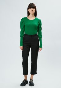 Grünes geripptes Langarmoberteil mit puffigen Schultern, kombiniert mit hoch taillierten schwarzen Hosen und klobigen schwarzen Loafers mit goldener Kettendetails.