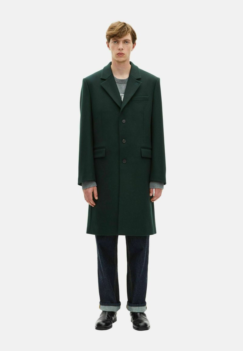 Abrigo de lana verde con solapas puntiagudas, botones frontales, dos bolsillos laterales y un bolsillo en el pecho, superpuesto a un suéter gris y unos jeans oscuros.