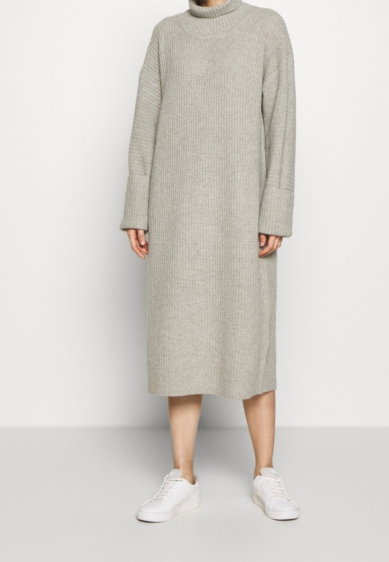Robe tricotée gris clair avec un col haut et des manches longues ; texture côtelée ; hauteur genou ; associée à des baskets blanches.