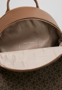 Tan round backpack with a textured exterior, gold zipper, and a beige interior featuring a repeated logo pattern.