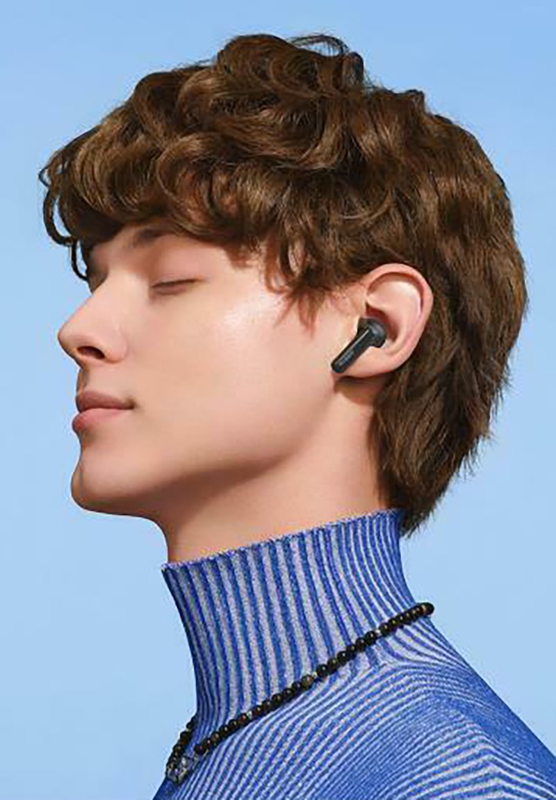 Wireless earbuds in a matte black finish, worn in a model’s ear, with a textured blue striped turtleneck and a black beaded necklace.