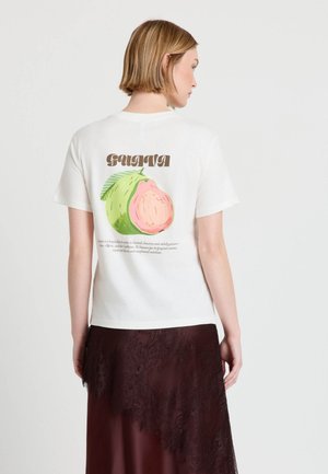 Donna con capelli corti biondi che indossa una maglietta bianca con un'illustrazione del frutto della guava e testo marrone sul retro, abbinata a una gonna di pizzo scuro.