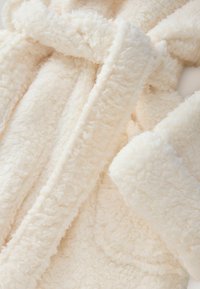 Robe de couleur crème en tissu doux et moelleux avec une surface texturée. Dotée d'un col châle et d'une ceinture à nouer.