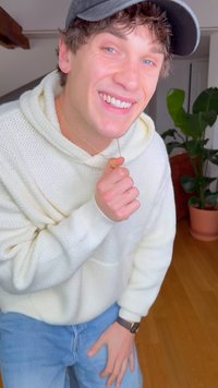 Felpa con cappuccio bianca lavorata a maglia con tasca, jeans azzurri e una collana d'oro. Sullo sfondo una pianta verde e pavimento in legno.