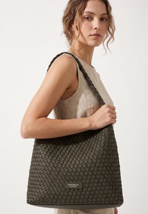 Vrouw met licht krullend haar die een mouwloze beige jurk draagt en een grote donkergroene geweven schoudertas met het label Diana&Co Firenze draagt.