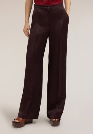 Femme portant un pantalon large marron foncé et des sandales compensées marron, une main dans la poche et un top rouge raccourci.