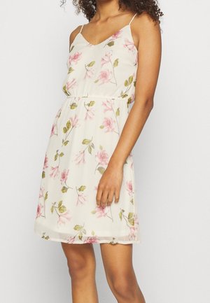 Kobieta ubrana w białą sukienkę bez rękawów z różowym kwiatowym wzorem i cienkimi ramiączkami, stojąca na jasnym, jednolitym tle.