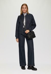 Dunkelblaues Jeansjackett mit einem Reißverschluss, grauer Rollkragenpullover darunter, weite dunkle Jeanshose und eine schwarze Umhängetasche. Schwarze klobige Schuhe.