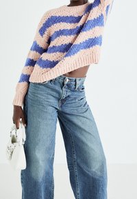 Personne portant un pull en maille rayé rose et bleu et un jean taille haute à jambes larges, tenant un petit sac à main blanc sur fond blanc.