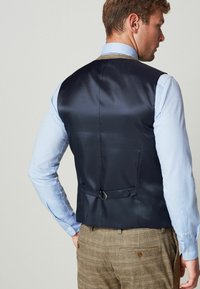 Muž v modrej košeli s dlhým rukávom, s námořníckou saténovou vestou so nastaviteľným pásom a hnedými kockovanými nohavicami, z pohľadu zozadu na neutrálnej pozadí.