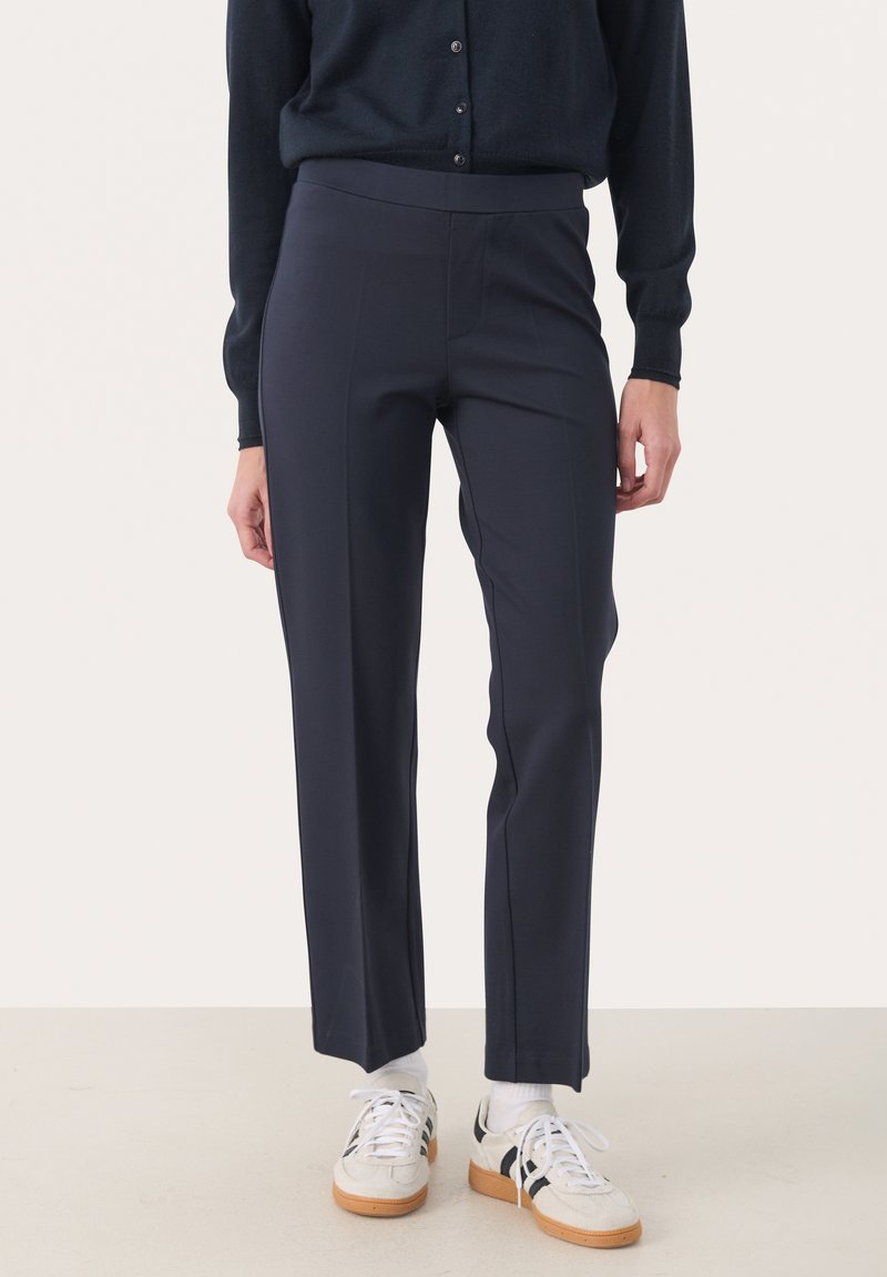 Pantalons ajustés bleu marine en tissu extensible, dotés d'une taille haute, de jambes droites et de coutures frontales discrètes. Associés à des baskets blanches.