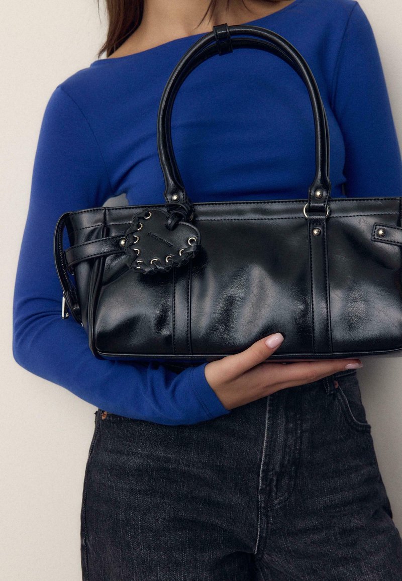 Femme vêtue d'un haut à manches longues bleu tenant un sac à main en cuir noir avec une étiquette en forme de cœur, portant un jean gris foncé sur un fond uni.