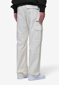 Lyse beige cargobukser med flere lommer, lett glans, bred ben-design, og synlige sømdetaljer, kombinert med hvite joggesko.