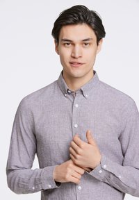 Camisa gris de botones con un tejido texturizado, de mangas largas y botones blancos, abotonada parcialmente en el cuello, usada por una persona.