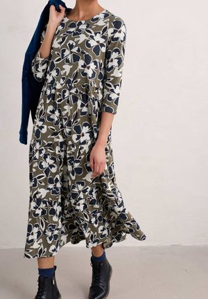 Femme portant une robe midi verte à fleurs avec manches 3/4, des bottines noires et tenant une veste bleu marine sur l'épaule.