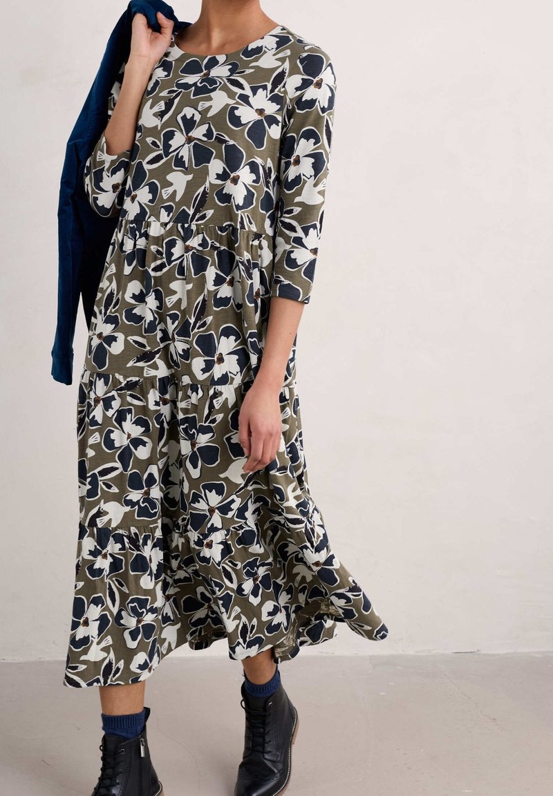 Femme portant une robe midi verte à fleurs avec manches 3/4, des bottines noires et tenant une veste bleu marine sur l'épaule.