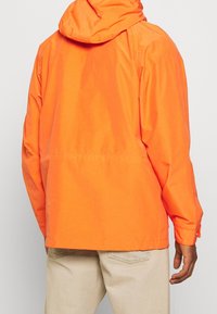 Oranžová bunda s kapucí, vyrobená z hladkého, lehkého materiálu. Design zahrnuje mírně volný střih a nastavitelnou manžetu.