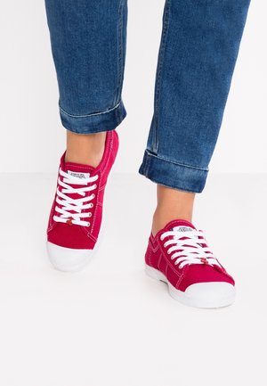 Osoba ubrana w podwinięte niebieskie dżinsy i jaskrawo czerwone trampki z materiału z białymi sznurówkami i gumowymi czubkami, stojąca na białej podłodze.