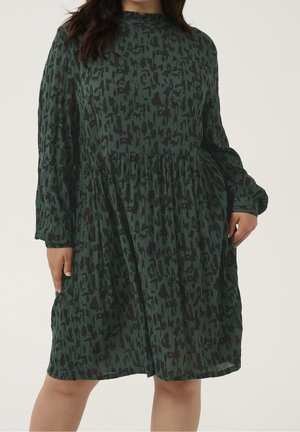 Donna che indossa un vestito verde a maniche lunghe, lungo fino al ginocchio, con un motivo astratto nero e una vita arricciata.