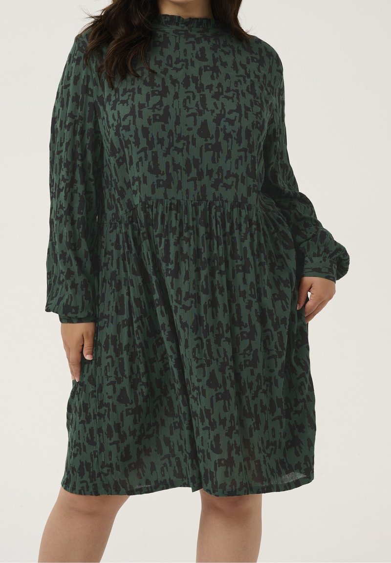 Femme portant une robe verte à manches longues, longueur genou, avec un motif abstrait noir et une taille froncée.