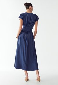 Temno modra maxi obleka s kratkimi rokavi, z gubanjem v pasu in tekočim krilom. Tkanina je videti gladka. Zadaj je majhna zadrga.