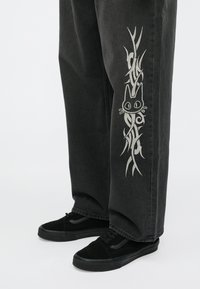 Svarta jeansbyxor med ett dekorativt ljusgrått kattryck och ett virvlande mönster på vänster ben, kombinerade med svarta sneakers.