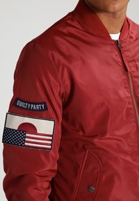 Czerwona nylonowa kurtka bomberka z prążkowanym kołnierzem, zamkiem błyskawicznym, boczną kieszenią oraz haftowanymi naszywkami amerykańskiej i japońskiej flagi.