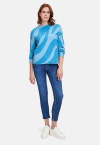 Blauer Pullover mit abstrakten, hellblauen Wellen, aus weichem Stoff. Kombiniert mit engen Jeans und weißen Sneakern, was einen lässigen Look schafft.
