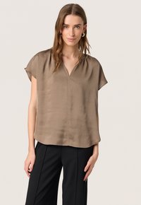 Blouse marron légère avec un décolleté en V, des manches courtes et une texture lisse. Assortie à un pantalon noir, elle offre une coupe décontractée.