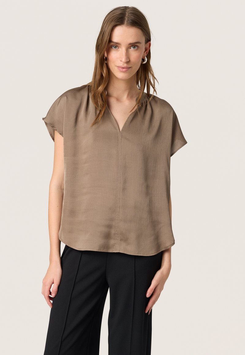Blouse marron légère avec un décolleté en V, des manches courtes et une texture lisse. Assortie à un pantalon noir, elle offre une coupe décontractée.