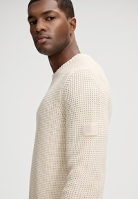 Jasnobeżowy sweter dziany z okrągłym dekoltem, teksturowanym wzorem oraz boczną metką z nazwą marki w formie wypukłego wzoru.