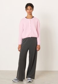 Žena stojící v růžovém zapínaném svetru, volných tmavě šedých kalhotách a šedých teniskách na světlém pozadí.