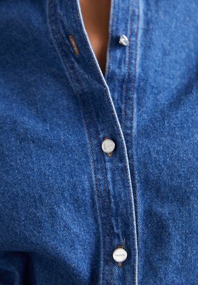 Denimskjorte med en mørkeblå farge og teksturert overflate, standard krage og sølvknapper, som fremhever et klassisk design.