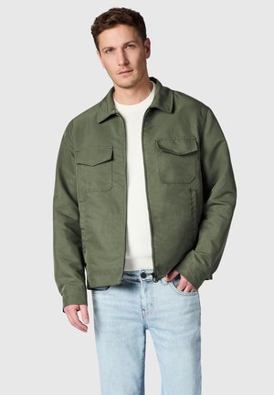 Homme portant une veste zippée vert olive sur un pull blanc, associée à un jean bleu clair, debout devant un fond gris uni.