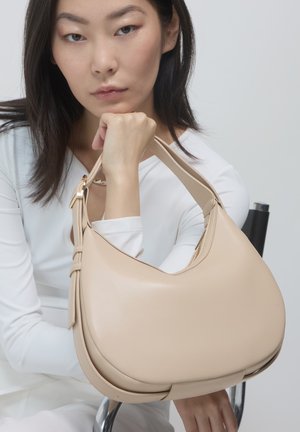 Beige Lederhandtasche mit einer geschwungenen Silhouette, einem Einzelriemen und sichtbaren Steppnähten. Glatte Textur und minimale Hardware-Akzente.