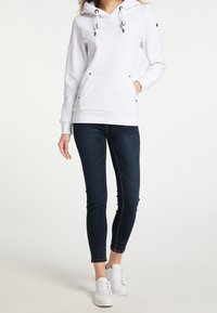 Sudadera blanca con un bolsillo canguro, cordones negros y ojales plateados, combinada con jeans skinny azul oscuro y zapatillas blancas.